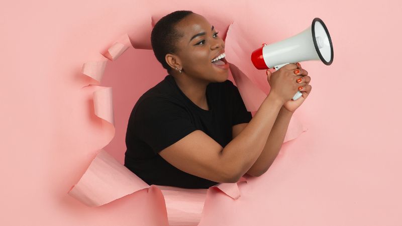 Black woman holding a megaphone after bursting through a pink paper background.